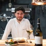 portrait de Quentin Maufrais souriant avec un verre de vin et une bouteille de Versant Viognier des Vignobles de Foncalieu