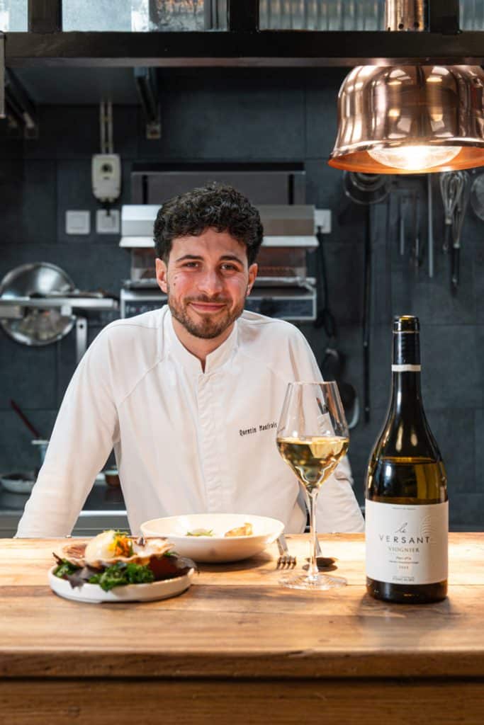 portrait de Quentin Maufrais souriant avec un verre de vin et une bouteille de Versant Viognier des Vignobles de Foncalieu